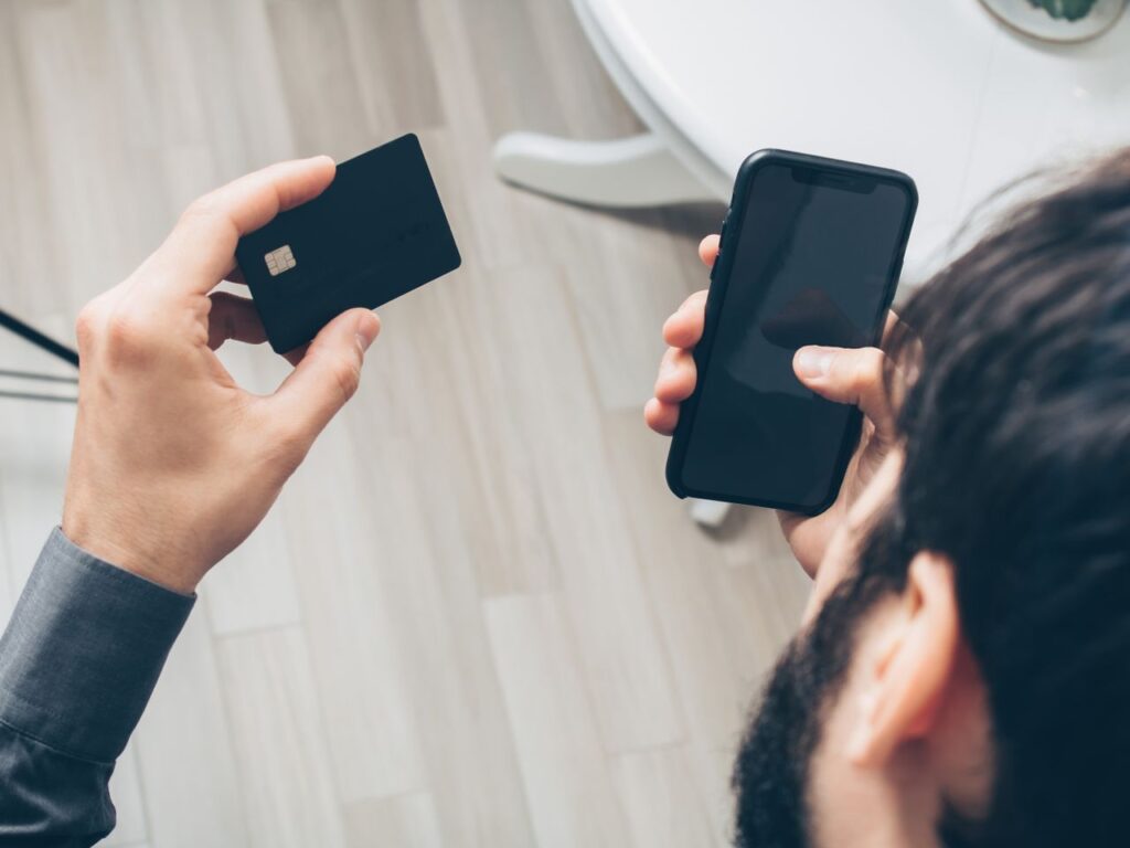 pexels-photo-7534379-7534379 Close-up of man holding a smartphone and credit card, making an online payment indoors.